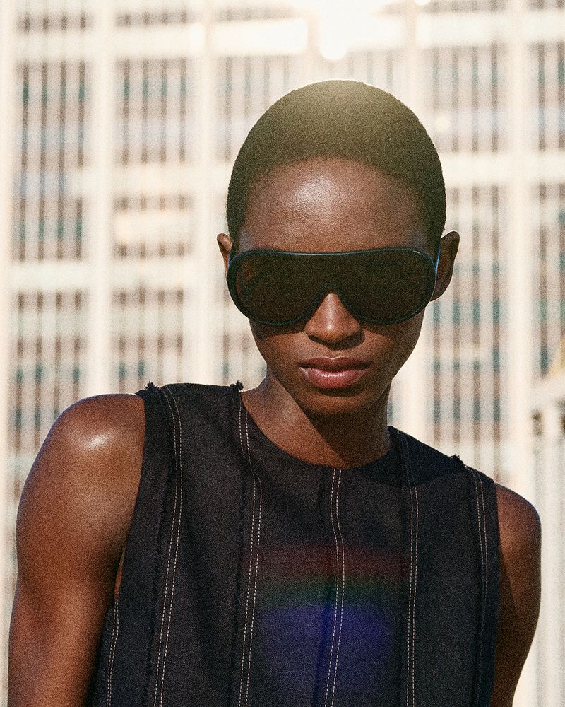 A dark-skinned woman wearing sunglasses and a dark sleeveless top stands against a neutral-toned building, bathed in bright sunlight.