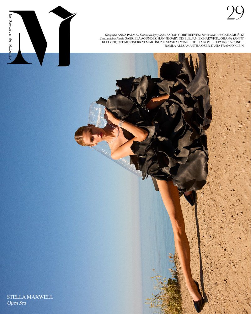 Model Stella Maxwell wearing a black ruffled dress reclining on a sandy beach against the ocean and blue sky.