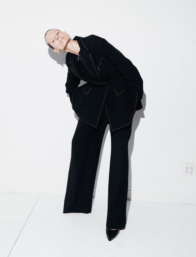 Woman in a black suit strikes a pose in a studio. The long-sleeved blazer and pants are accented with white stitching detail.