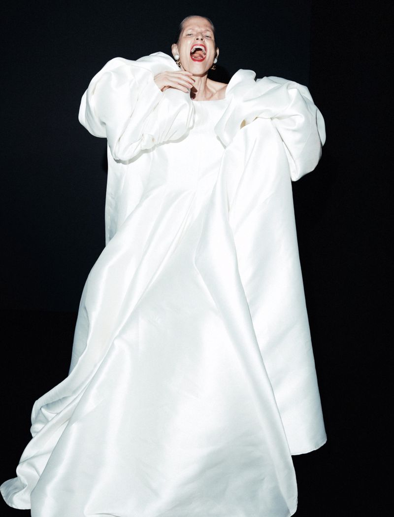 A woman in a dramatic white gown with large sleeves stands against a black background, captured in a striking fashion-style pose.