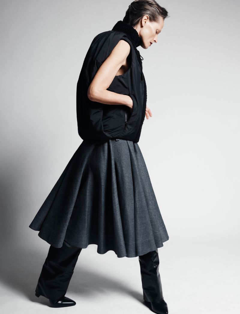 Fashionable adult woman posing in studio wearing a black puffer vest, gray skirt, black pants, and high black boots on white backdrop.