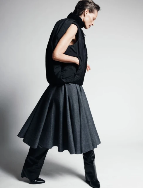 Fashionable adult woman posing in studio wearing a black puffer vest, gray skirt, black pants, and high black boots on white backdrop.