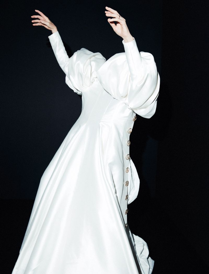 Elegant, surreal fashion image of a person in a couture gown with puffy sleeves against a stark black backdrop, minimal styling.