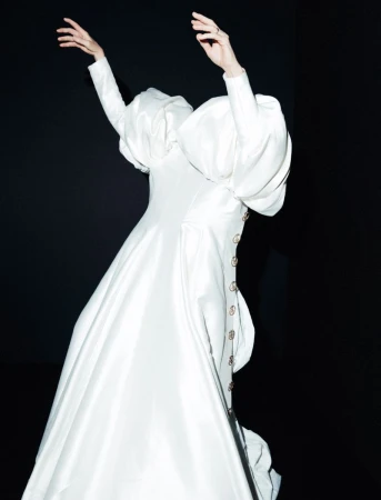 Elegant, surreal fashion image of a person in a couture gown with puffy sleeves against a stark black backdrop, minimal styling.