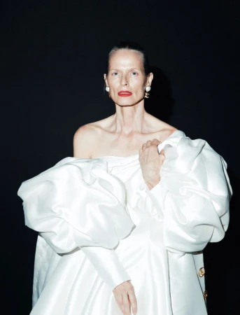 Elegant older woman poses in a white dress with puffy sleeves against a dark background, accessorized with red lipstick and pearl earrings.