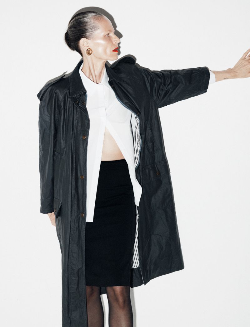 A woman in a black coat and skirt stands against a white background, angled three-quarters, with her arm extending to the right.