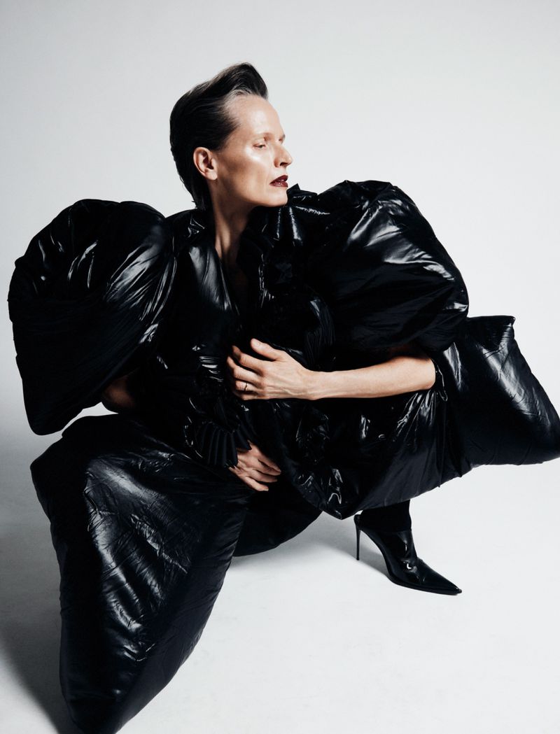 A woman in a glossy black puffer coat and heels poses in a studio, eyes closed, against a white background. Fashion editorial shot.