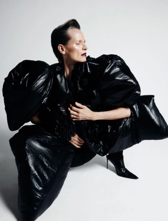 A woman in a glossy black puffer coat and heels poses in a studio, eyes closed, against a white background. Fashion editorial shot.
