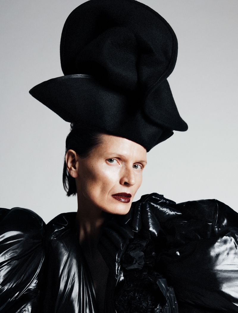 Close-up portrait of an adult woman wearing a large black hat and an oversized, glossy black jacket against a neutral studio backdrop.