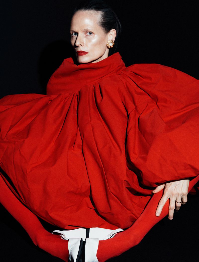 Elegant woman in a dramatic, oversized red gown with a high neckline, slicked-back hair, and red lipstick against a dark background.