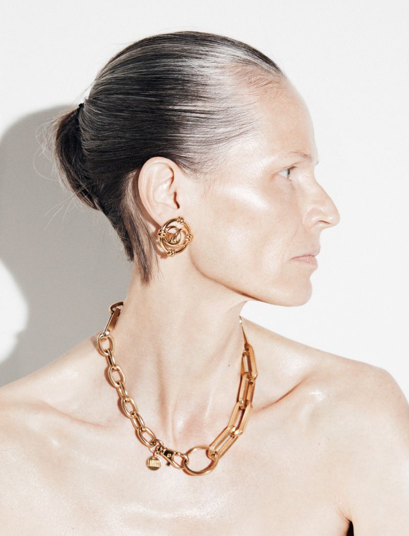 Profile portrait of model Sylke Golding wearing a bold gold link chain necklace and gold earring against a white background.