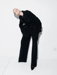 Model Sylke Golding poses leaning sideways wearing a tailored black pantsuit with white contrast stitching against a white wall.