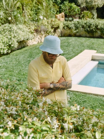 Man wearing a yellow camp shirt and light blue bucket hat standing near a pool with lush garden foliage