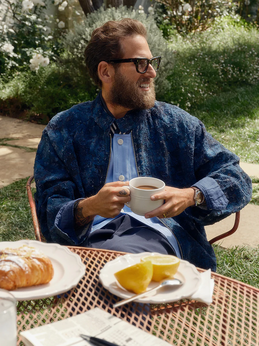 Jonah Hill wearing dark glasses and a blue jacket holding a white mug at an outdoor table with pastries