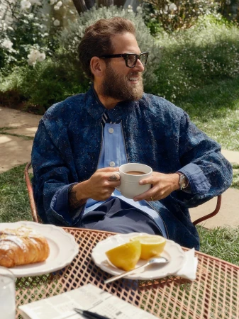 Jonah Hill wearing dark glasses and a blue jacket holding a white mug at an outdoor table with pastries