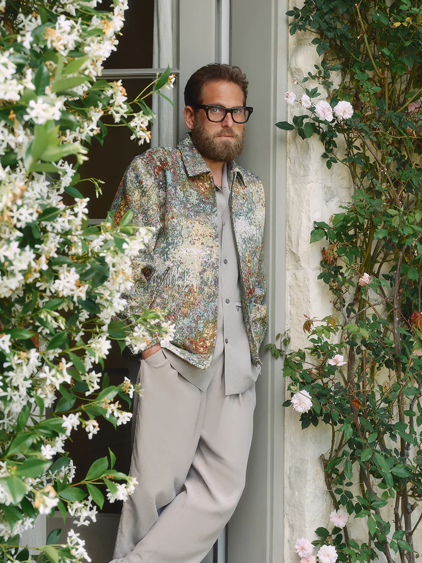 Jonah Hill wearing a patterned jacket and grey trousers standing in a doorway framed by flowering vines.