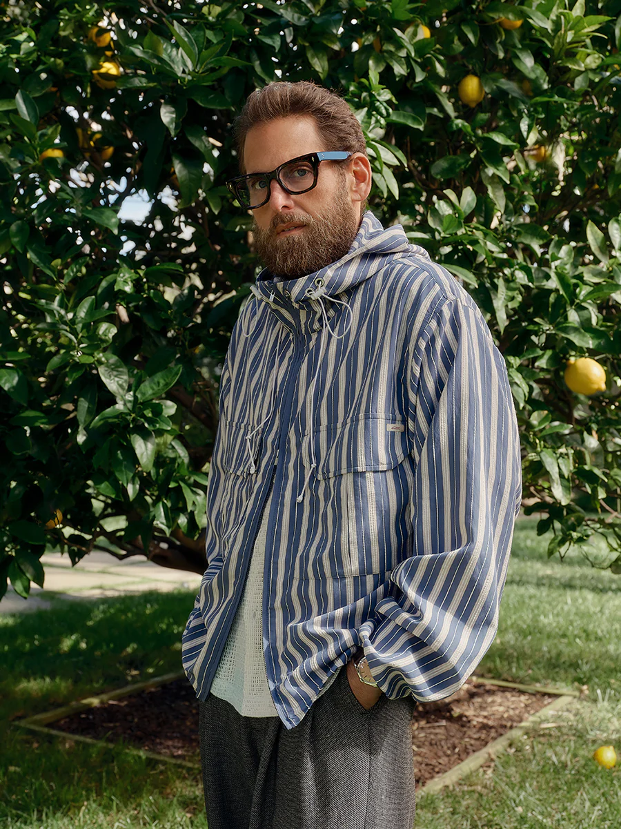 Jonah Hill wearing a blue and white striped hooded jacket and black glasses standing outdoors in front of a lemon tree.