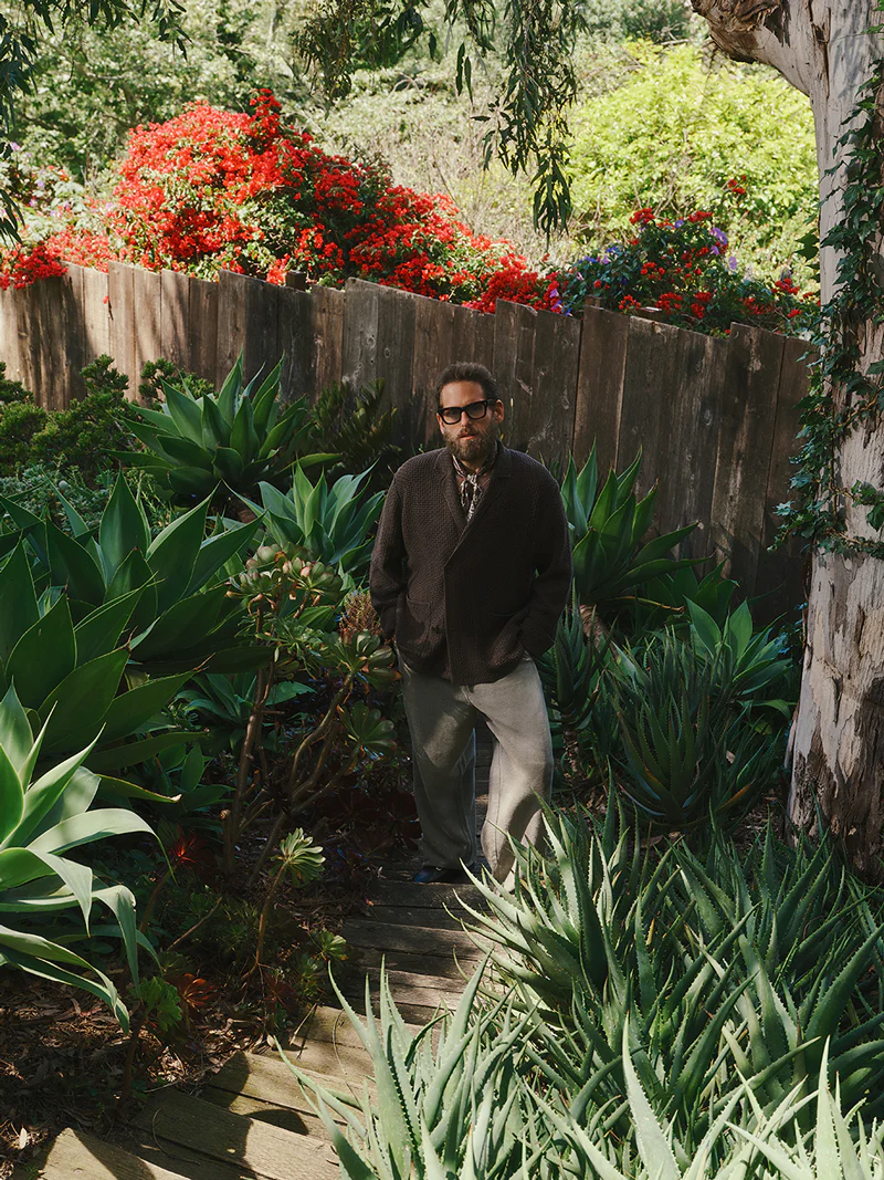 Man wearing sunglasses and a dark cardigan standing among lush green succulent plants outdoors