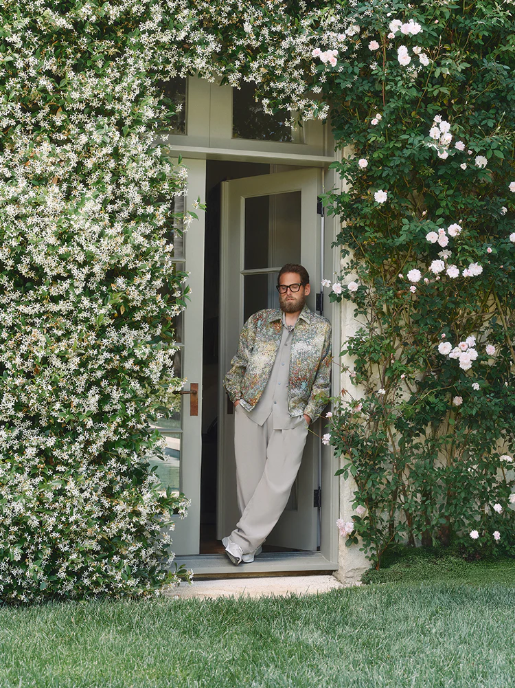 Jonah Hill leaning in a doorway framed by flowering vines, wearing a patterned jacket and light trousers for Kith.