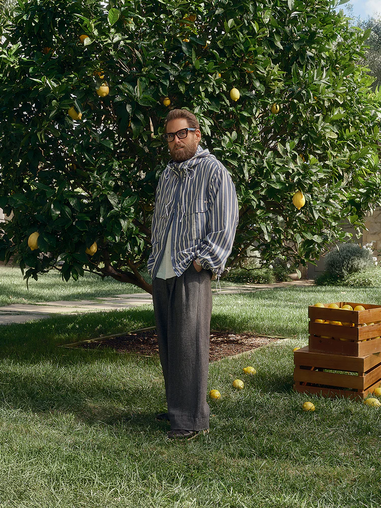 Jonah Hill wearing a striped jacket and glasses standing under a lemon tree in a grassy garden for Kith.