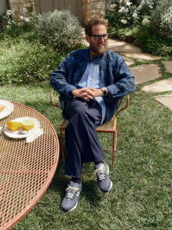 Jonah Hill seated in a wire chair outdoors wearing a blue jacket, button-down shirt, dark trousers, and sneakers