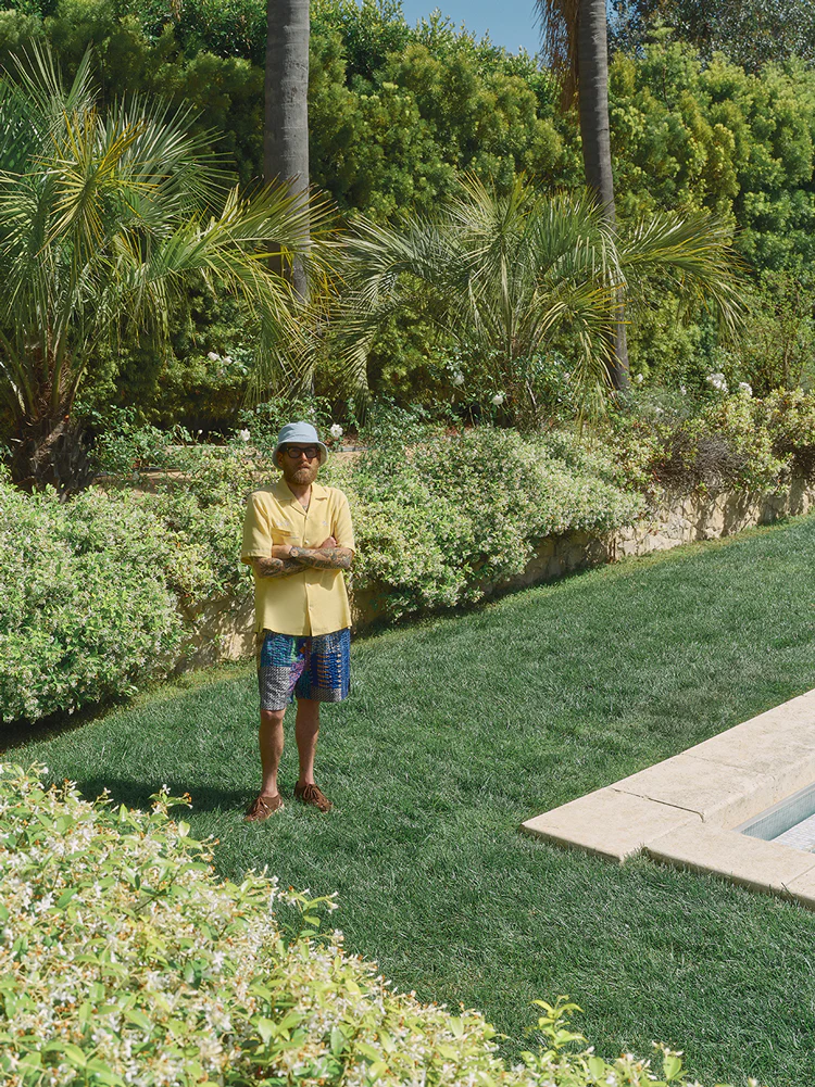 Man wearing a yellow shirt and patterned shorts standing on a lawn near a pool with palm trees in the background