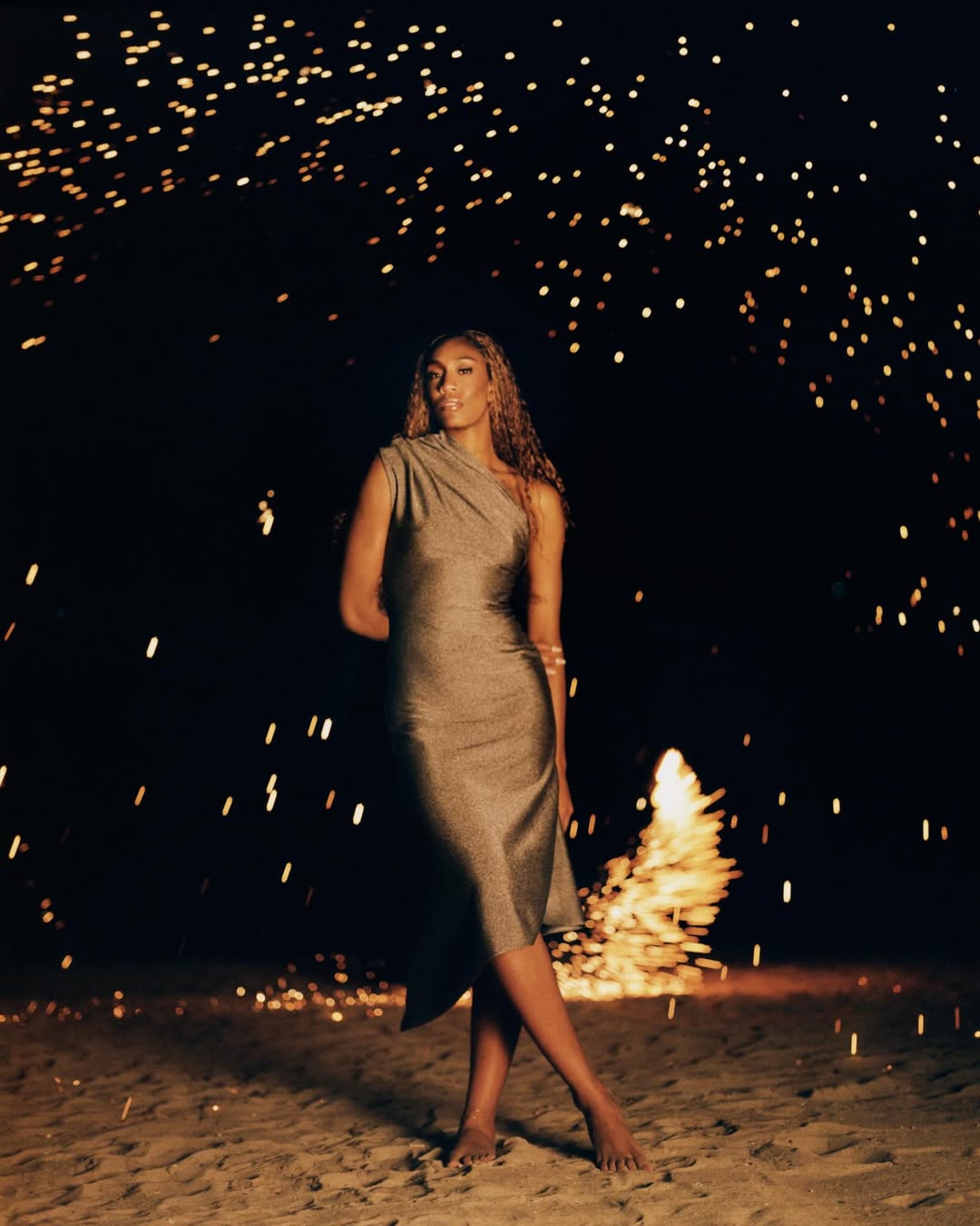A'ja Wilson in an asymmetric grey metallic dress standing on sand against a dark night background with glowing sparks.