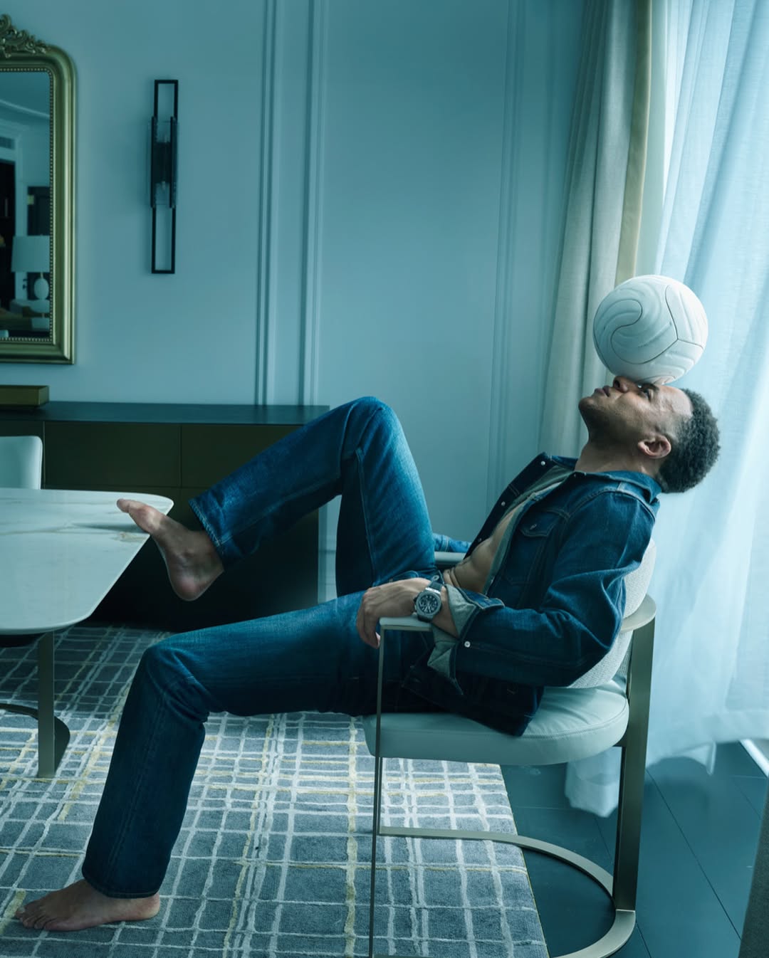 Kylian Mbappe seated backwards on a white chair balancing a white soccer ball on his forehead in a luxury interior.