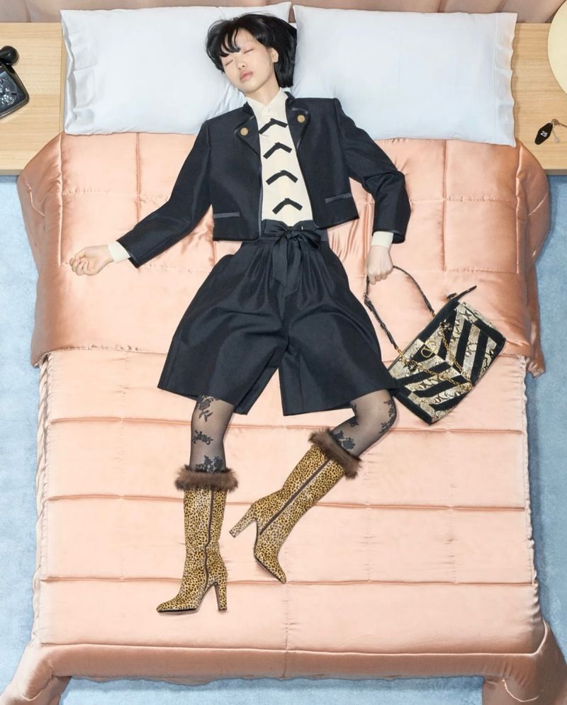 Woman in fashionable black jacket, shorts, and leopard-print boots lays on pink bed with patterned handbag in an indoor setting.