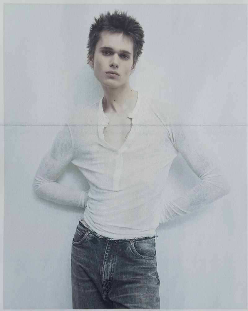 Young model with spiky hair wearing a white henley shirt and denim jeans against a pale grey seamless background.