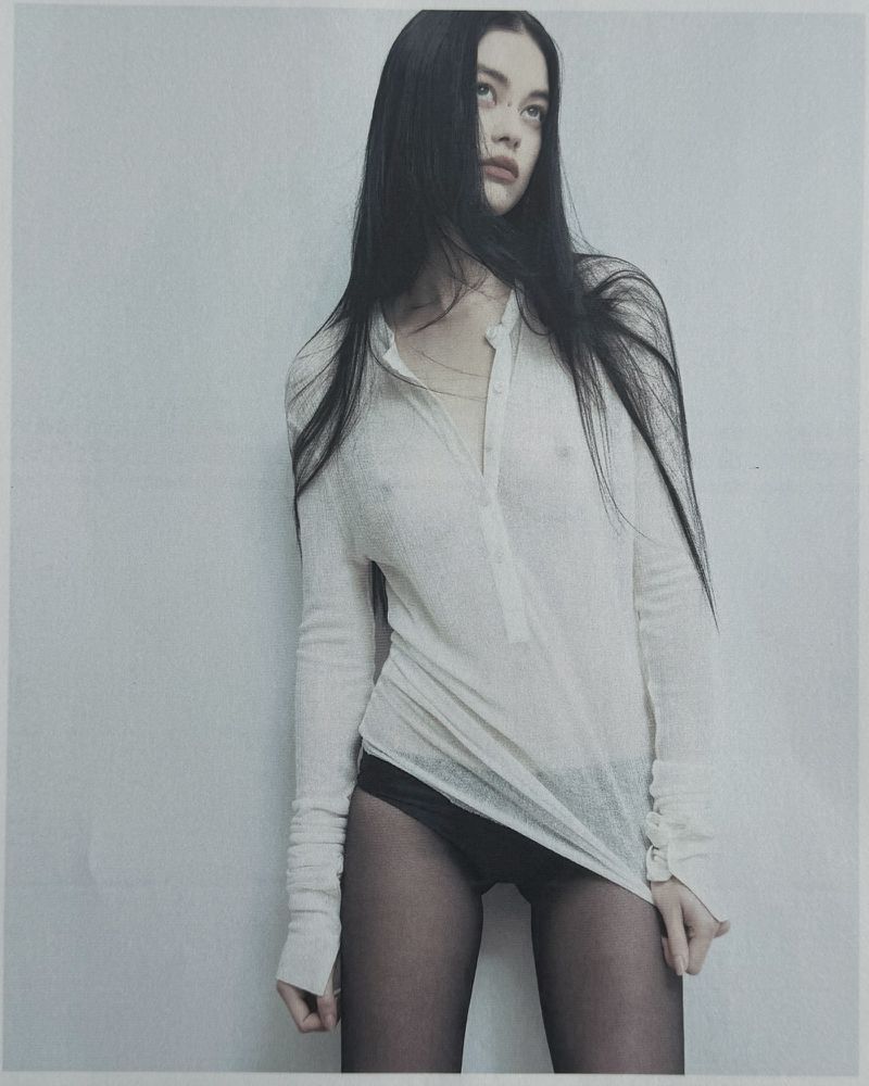 Brunette woman poses in a sheer top and black bottoms against a light grey wall. Studio lighting, fashion forward, close-up.