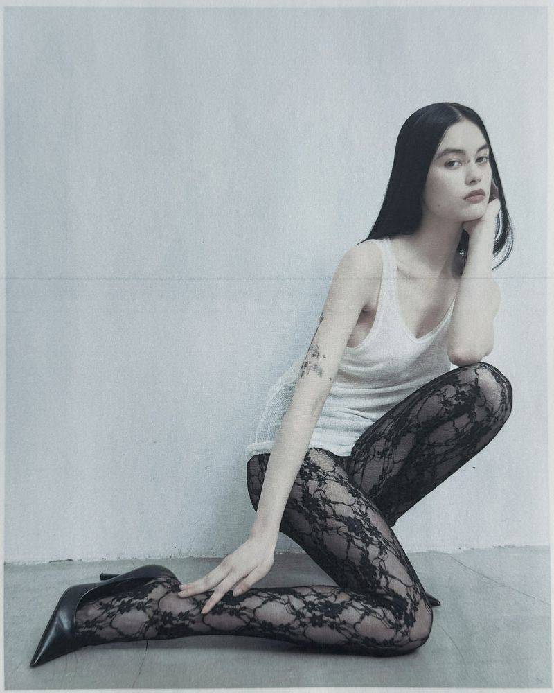 Model Mimi Dey crouching against a neutral wall in a white tank top and black lace tights with black heels for Unvain Studios SS26.