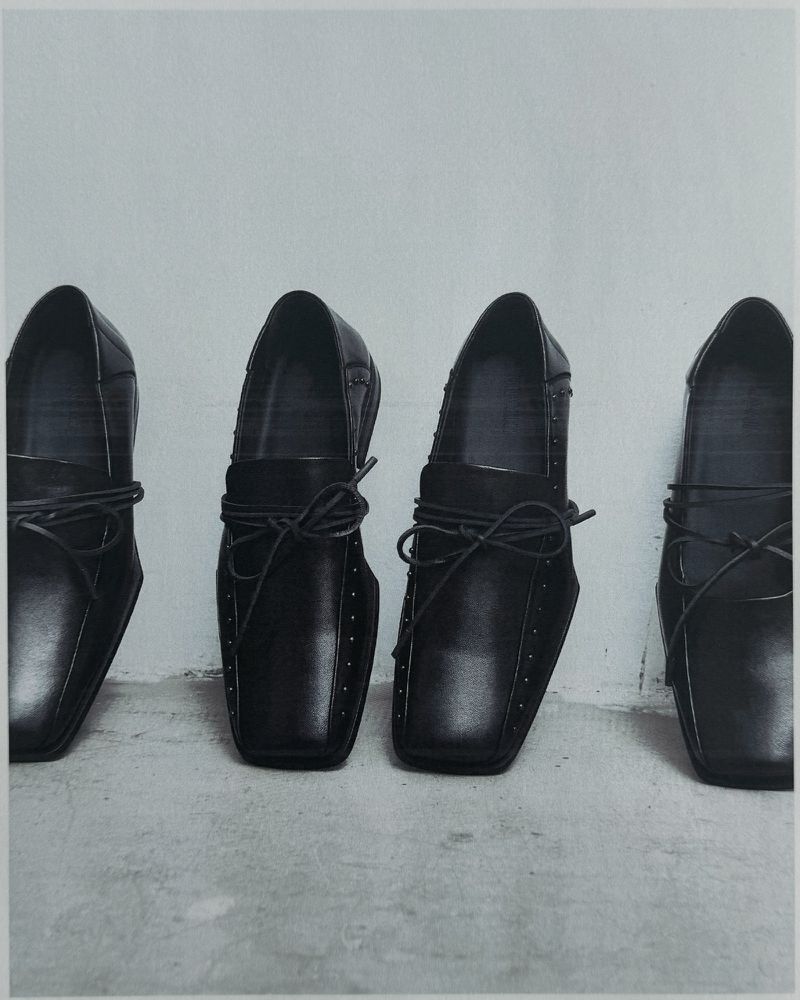 Four black leather loafers aligned in a row on a light grey concrete surface against a white wall.