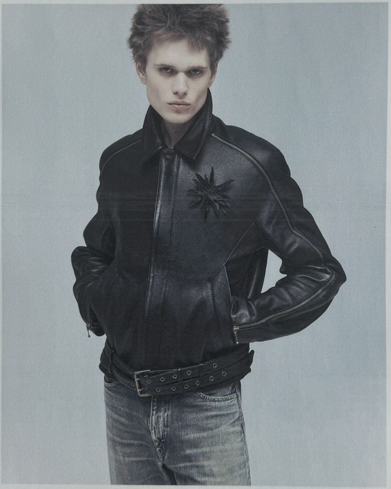 Model in black leather jacket and blue jeans posing against a plain grey seamless studio background.