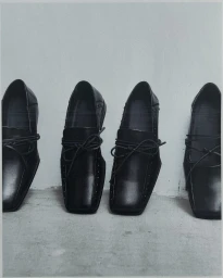 Four black leather loafers aligned in a row on a light grey concrete surface against a white wall.