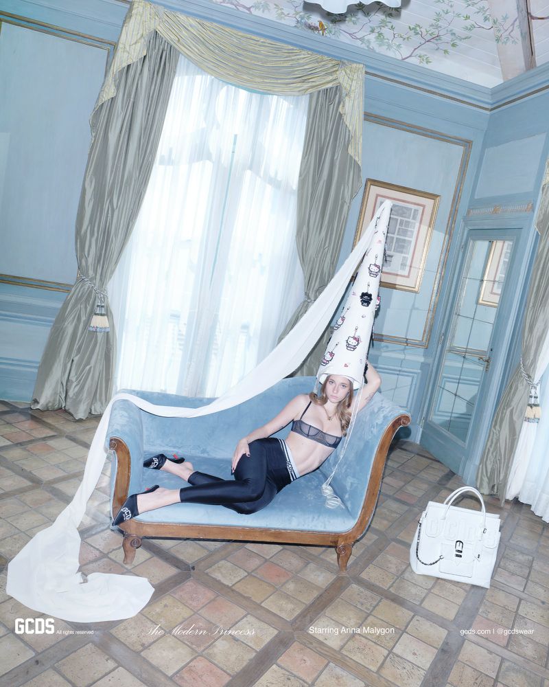 Model wearing tall conical hat and black lingerie set reclining on blue sofa in ornate room for GCDS campaign