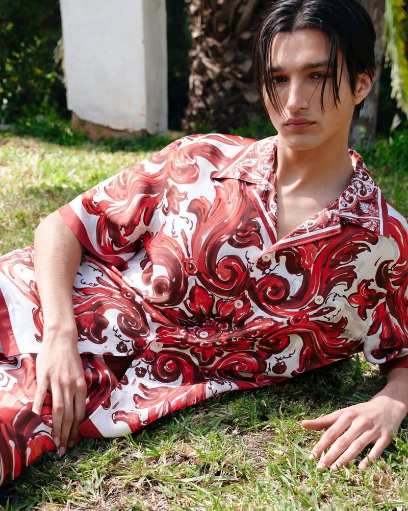 Man in patterned shirt lies on green grass outdoors in a medium shot, with fair skin and dark hair.