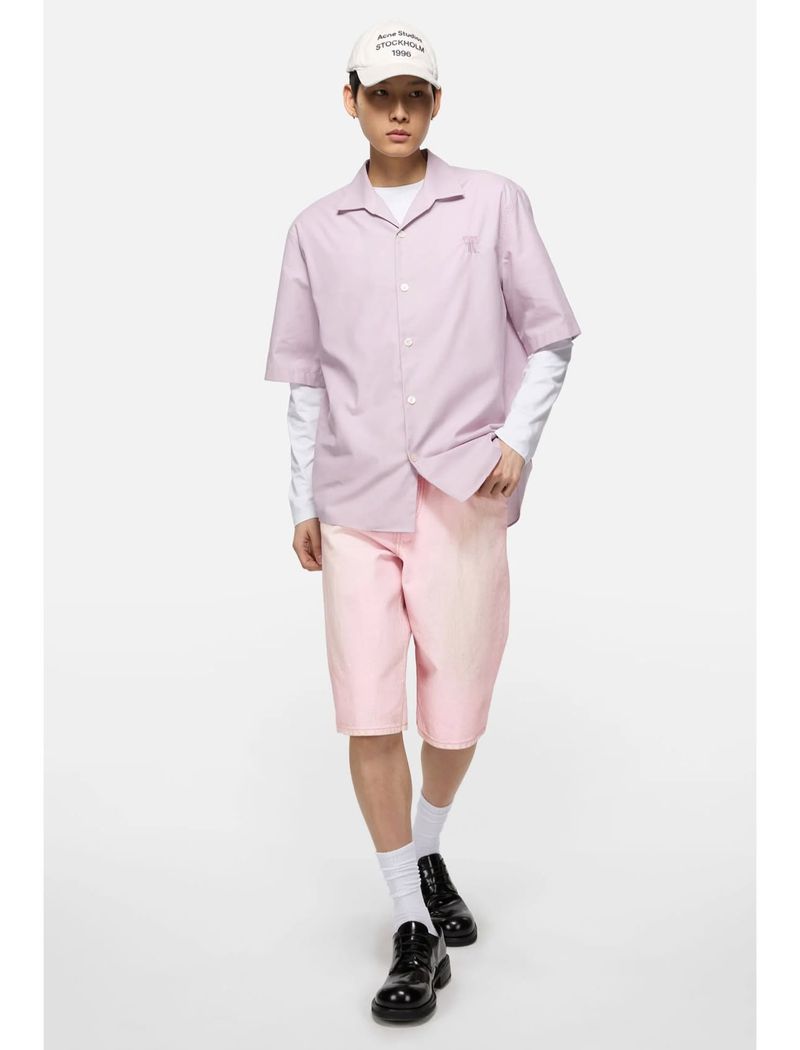 Fashion model posing in a lavender short-sleeved button-down shirt, pink shorts, white socks, and black dress shoes against white background.