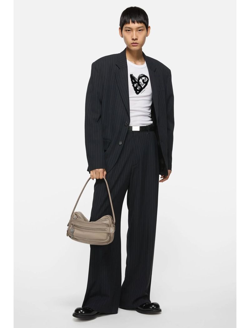 Young adult wearing oversized pinstripe suit, white t-shirt with graphic print, black belt and shoes and holding taupe handbag against white.