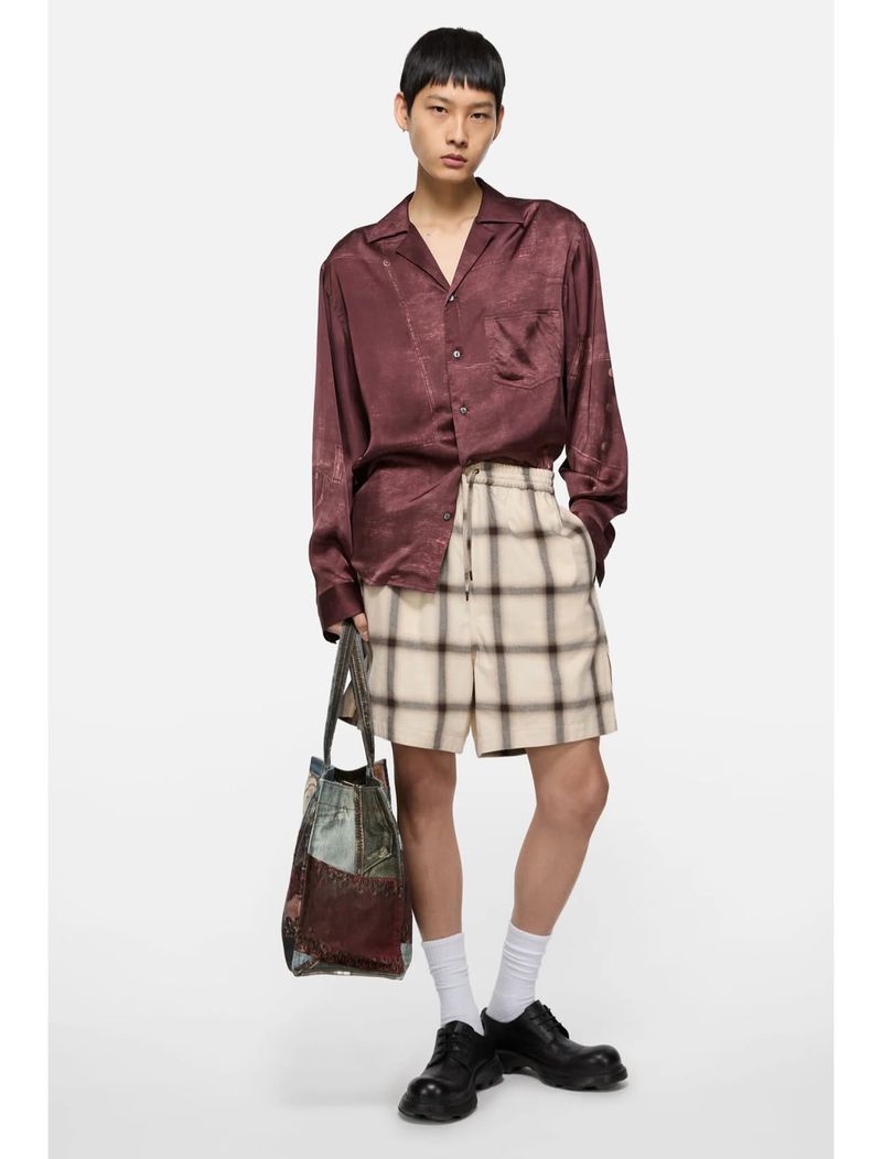 Male model in a maroon satin shirt and plaid shorts holds a tote bag against a bright background in a fashion editorial studio shot.