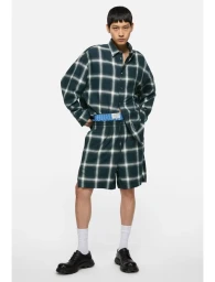 Model in dark green plaid shirt and matching shorts wearing white socks and black shoes against a white background.