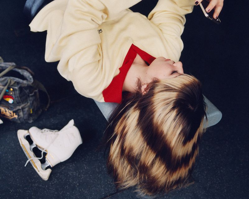 Close-up portrait of Alysa Liu lying back with highlighted hair spilling downward, wearing a cream sweater with a red collar