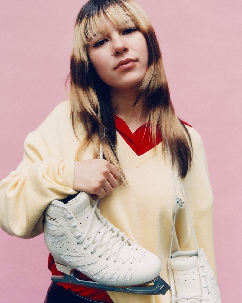 Alysa Liu holding white ice skates wearing a cream sweater against a pink background for W Magazine