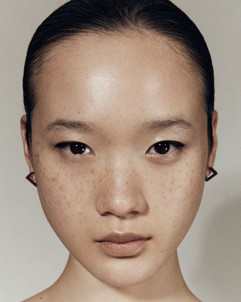 Close-up portrait of an adult woman with black eyeliner and minimalist square earrings against a plain background.