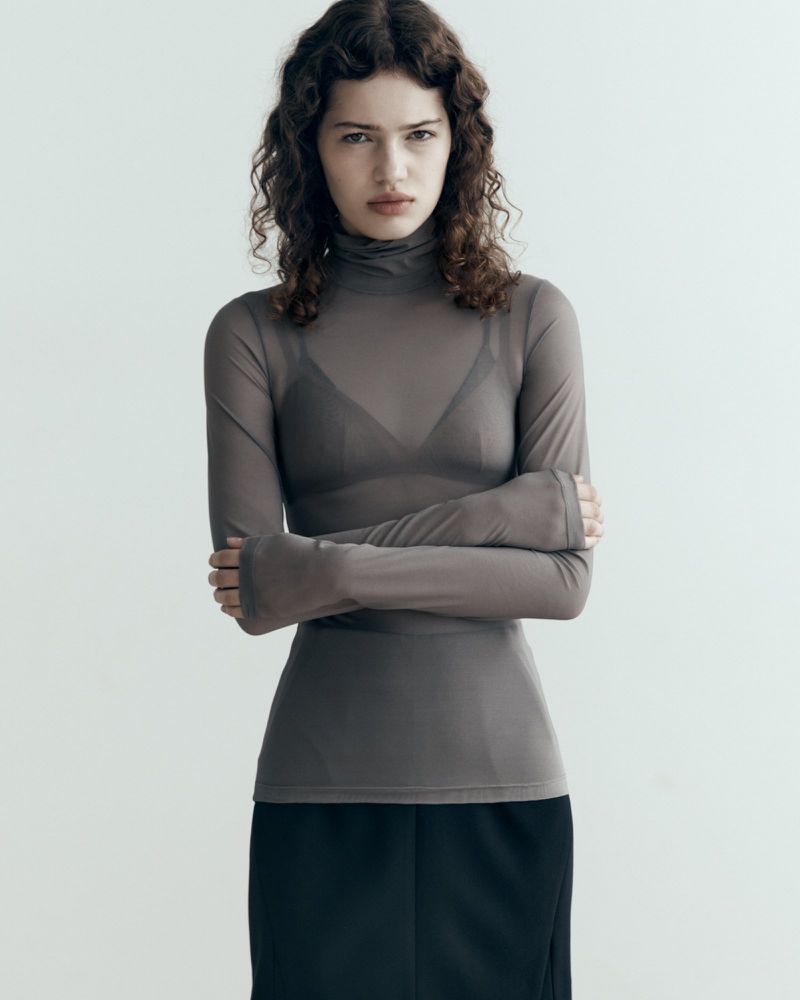 Fashion portrait of a woman with curly hair wearing a sheer grey turtleneck, black skirt, arms crossed, and facing forward in a studio.