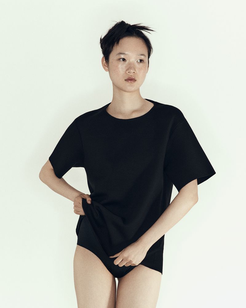 Woman in a black t-shirt and briefs stands indoors against a white backdrop in a studio portrait with a shallow depth of field.
