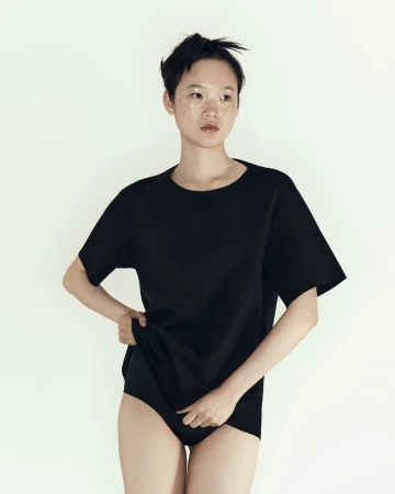 Woman in a black t-shirt and briefs stands indoors against a white backdrop in a studio portrait with a shallow depth of field.