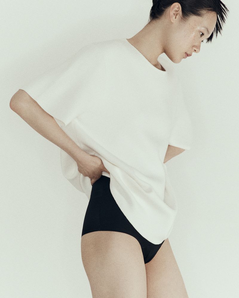 Asian woman poses in a photo studio wearing a white oversized t-shirt and black underwear against a neutral backdrop with soft lighting.