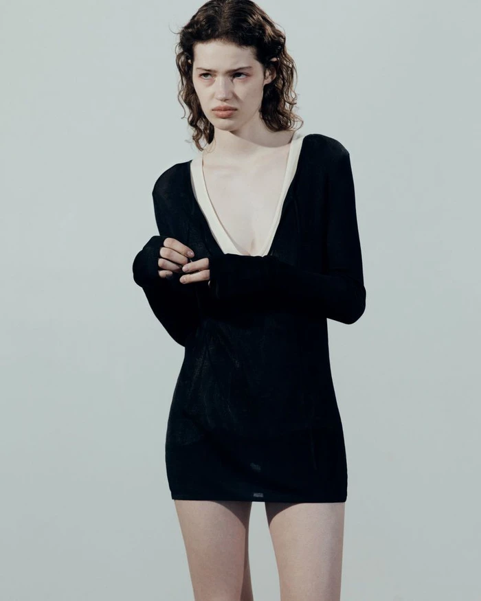 Model Nora van der Tuin wearing a black long-sleeve mini dress with a deep V-neck against a plain pale backdrop.