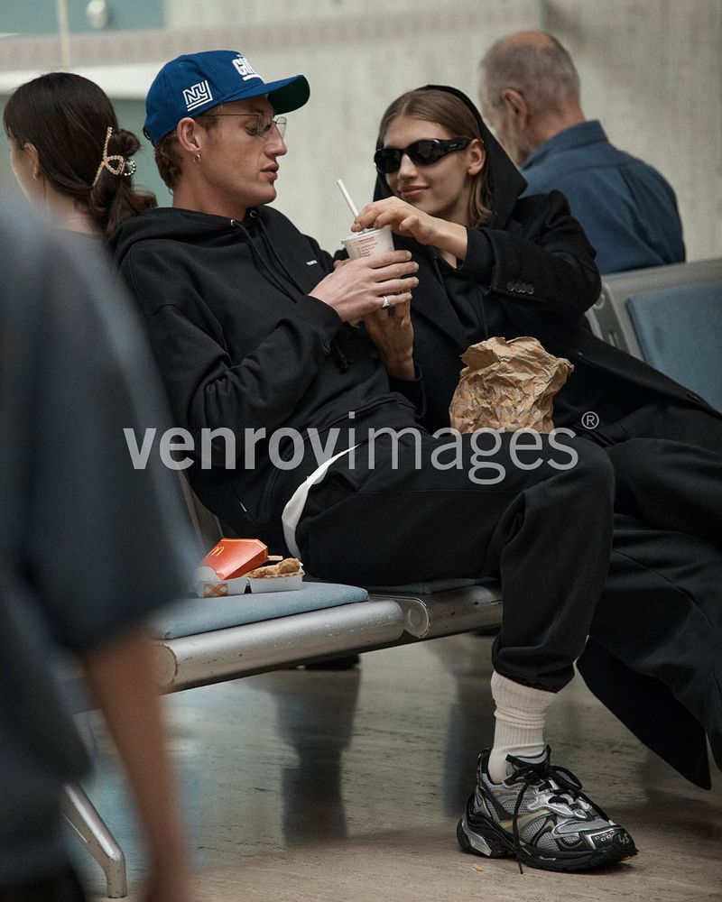 Two adults at an airport sit on a bench with a drink and snack. The woman wears sunglasses and a long coat, and the man a baseball cap.